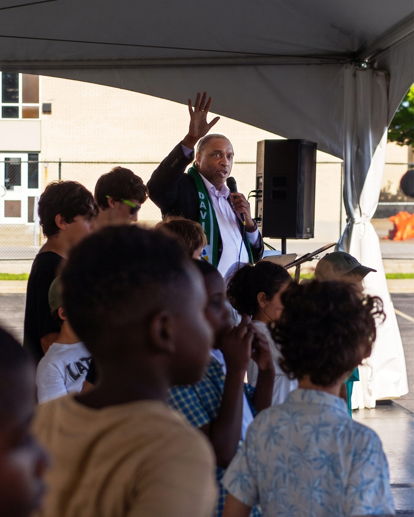 David Elisma au micro sous un chapiteau à Vanier, la main levée, entouré d'enfants rassemblés devant lui lors du Festi-Mosaïque.